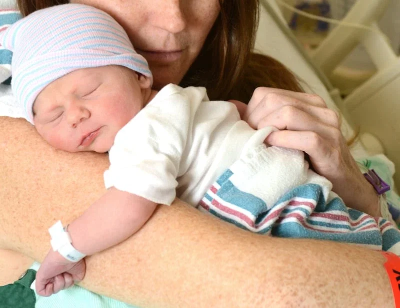 Mother holding sleeping newborn in hospital, infant wearing striped hat after birth injury delivery
