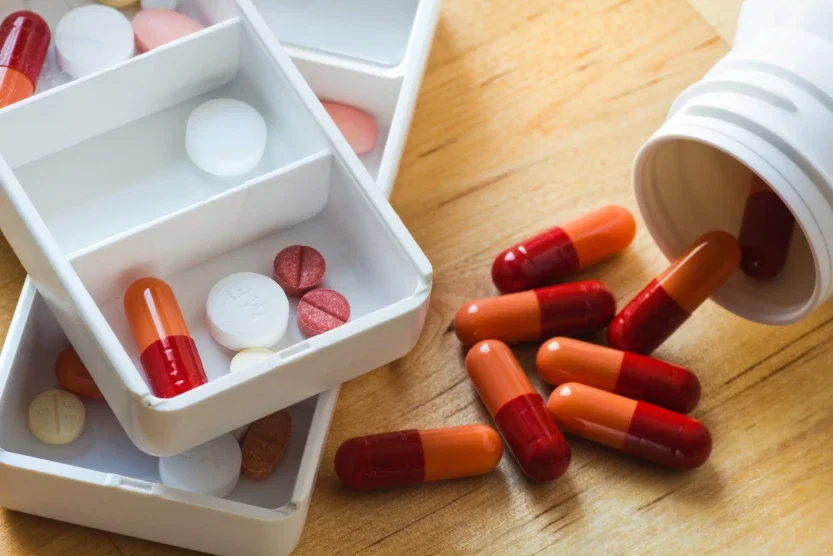 Assorted prescription medications in a weekly pill organizer alongside spilled capsules from a medication bottle