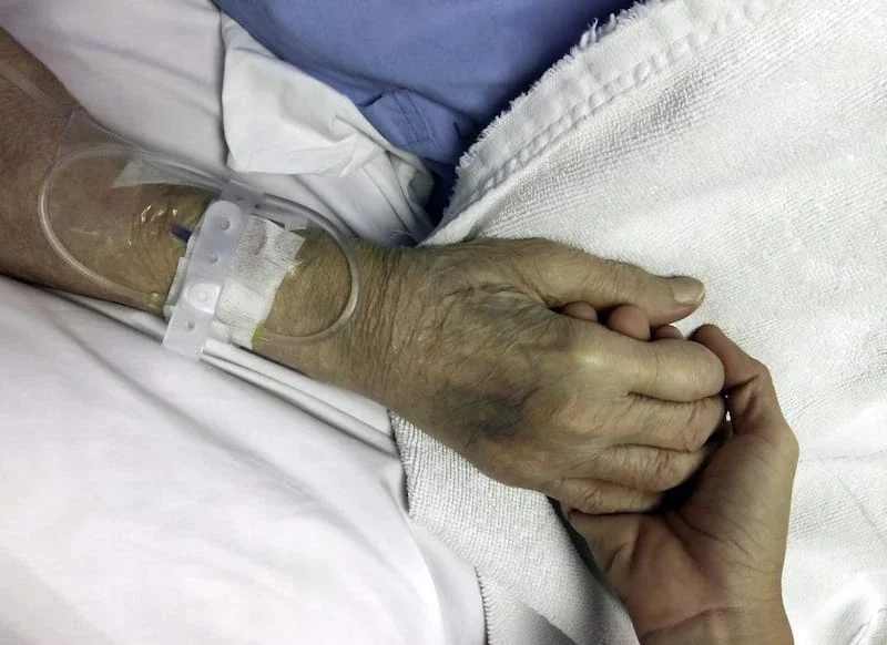 Young hand holding the frail elderly hand of a hospital patient with IV line attached
