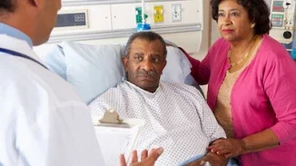 Doctor delivering difficult medical diagnosis to elderly male patient in hospital bed with wife at bedside