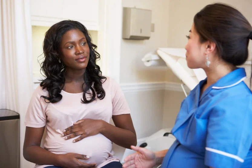 A pregnant woman sitting in a medical exam room with her hand on her belly, listening attentively to a nurse in blue scrubs