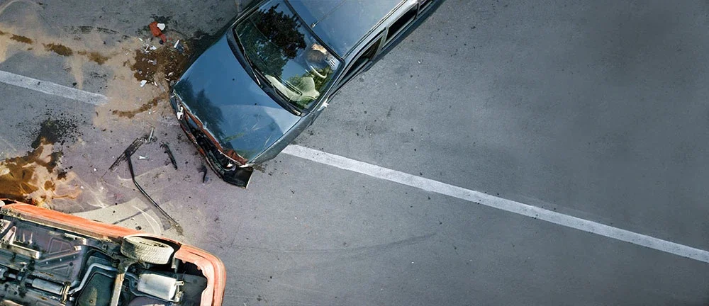 Aerial view of a two-vehicle accident on a road, showing a dark sedan with front-end damage and an overturned orange vehicle with fluid pooled on the pavement
