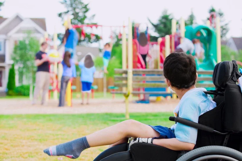 Child with cerebral palsy in wheelchair watching peers play at playground due to birth injury