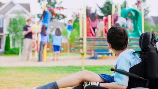 Child with cerebral palsy in wheelchair watching peers play at playground due to birth injury