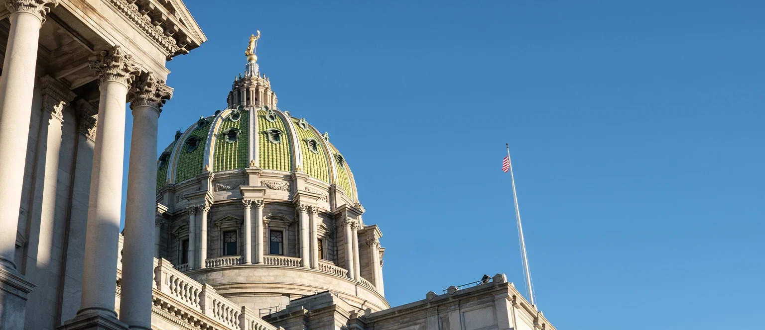 The green-tiled dome and ornate stone columns of the Pennsylvania State Capitol building in Harrisburg, with an American flag on a flagpole against a clear blue sky