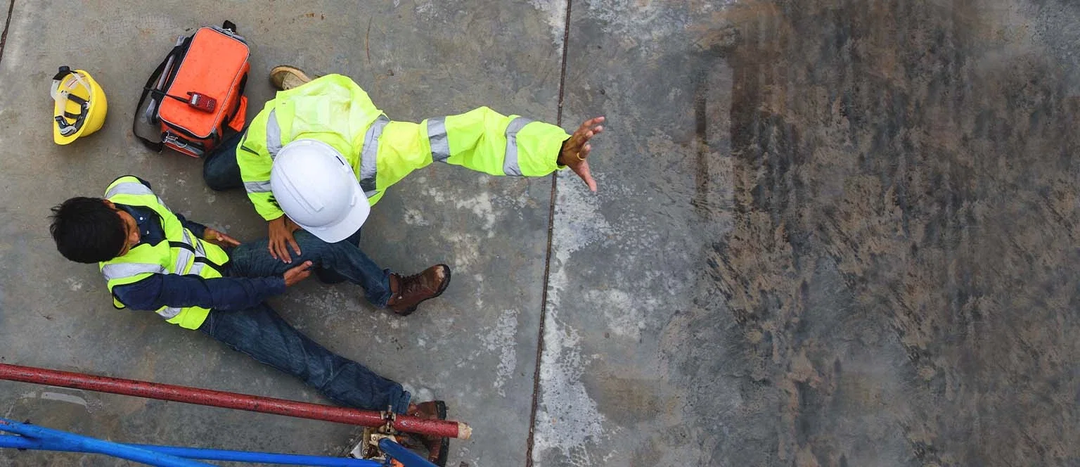Overhead view of a construction worker in a high-visibility vest kneeling to assist an injured coworker lying on a concrete surface, with a hard hat and first aid bag nearby