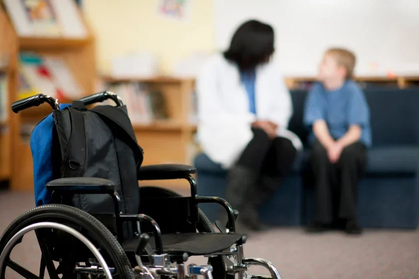 Empty wheelchair in foreground with child consulting doctor in medical office background