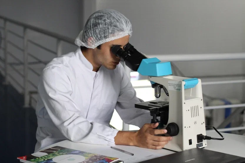 Lab technician in sterile cap examining specimen under microscope during medical research or testing