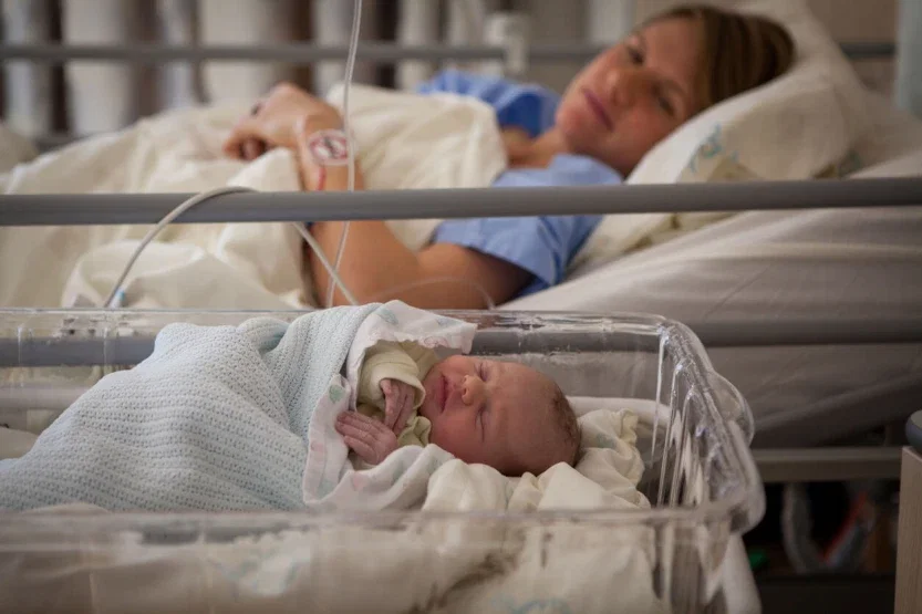 Newborn baby in hospital bassinet with mother resting in bed after labor and delivery