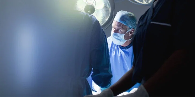 Surgeon in blue scrubs and mask looking up during operation under bright surgical lamp in darkened OR