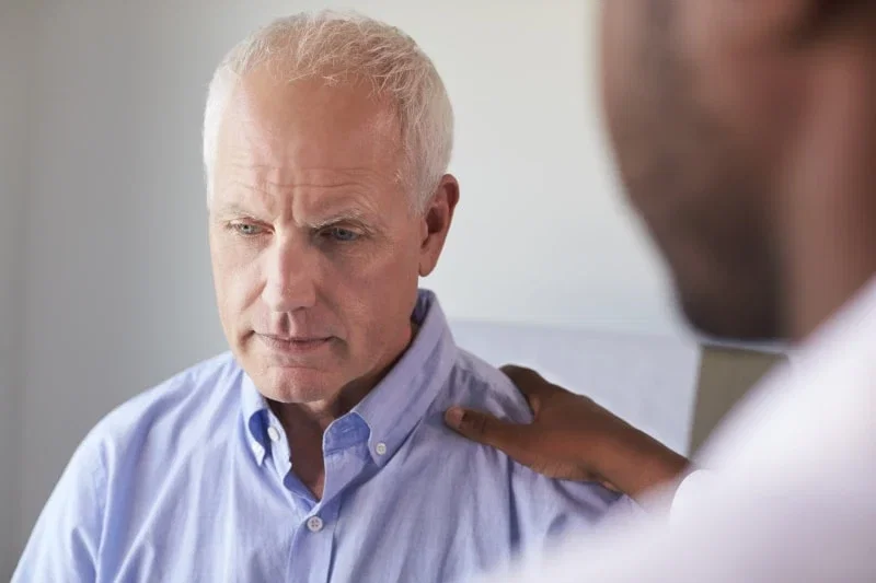 An older man with a concerned expression looking downward while a doctor places a supportive hand on his shoulder