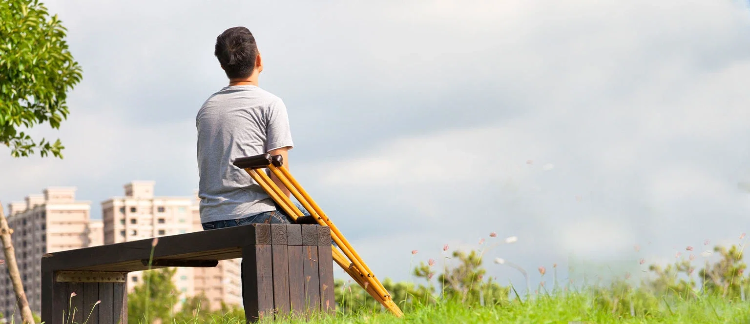A man with crutches resting beside him sitting alone on an outdoor bench, viewed from behind, looking out toward a cloudy sky and city buildings