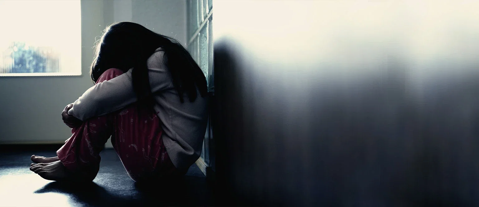 A young child sitting alone on a dark floor against a wall, hugging their knees to their chest in a dimly lit room