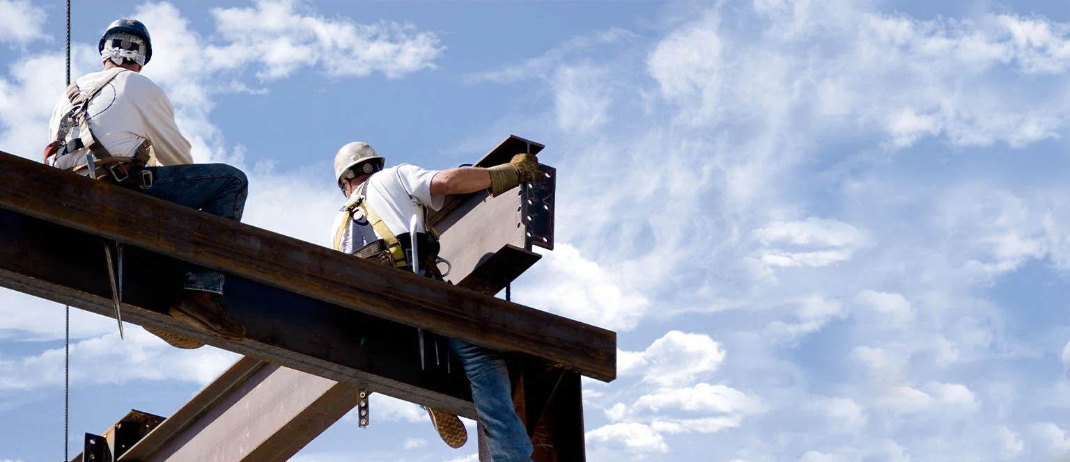 Two construction workers in hard hats and safety harnesses working high above the ground on steel I-beams against a partly cloudy sky