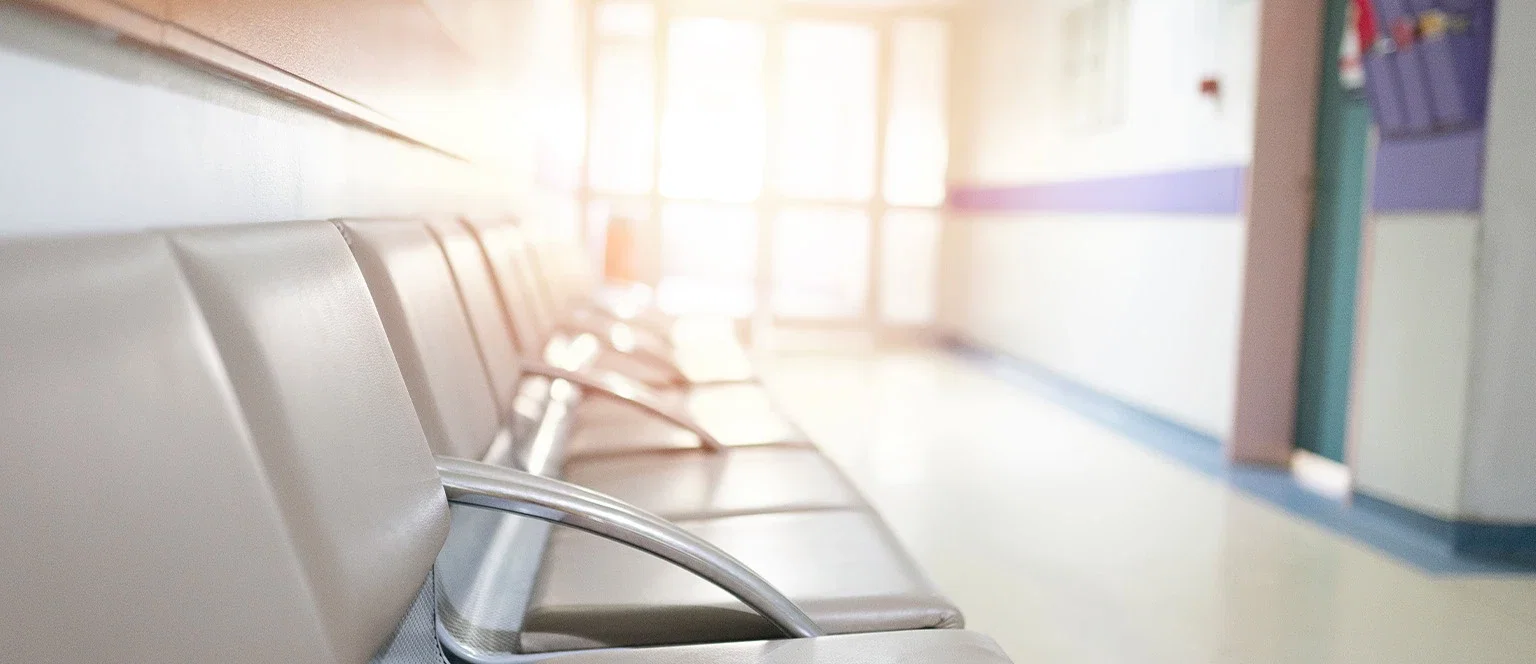 Empty row of beige waiting room chairs along a hospital corridor, with a blurred hallway and patient room door visible in the background