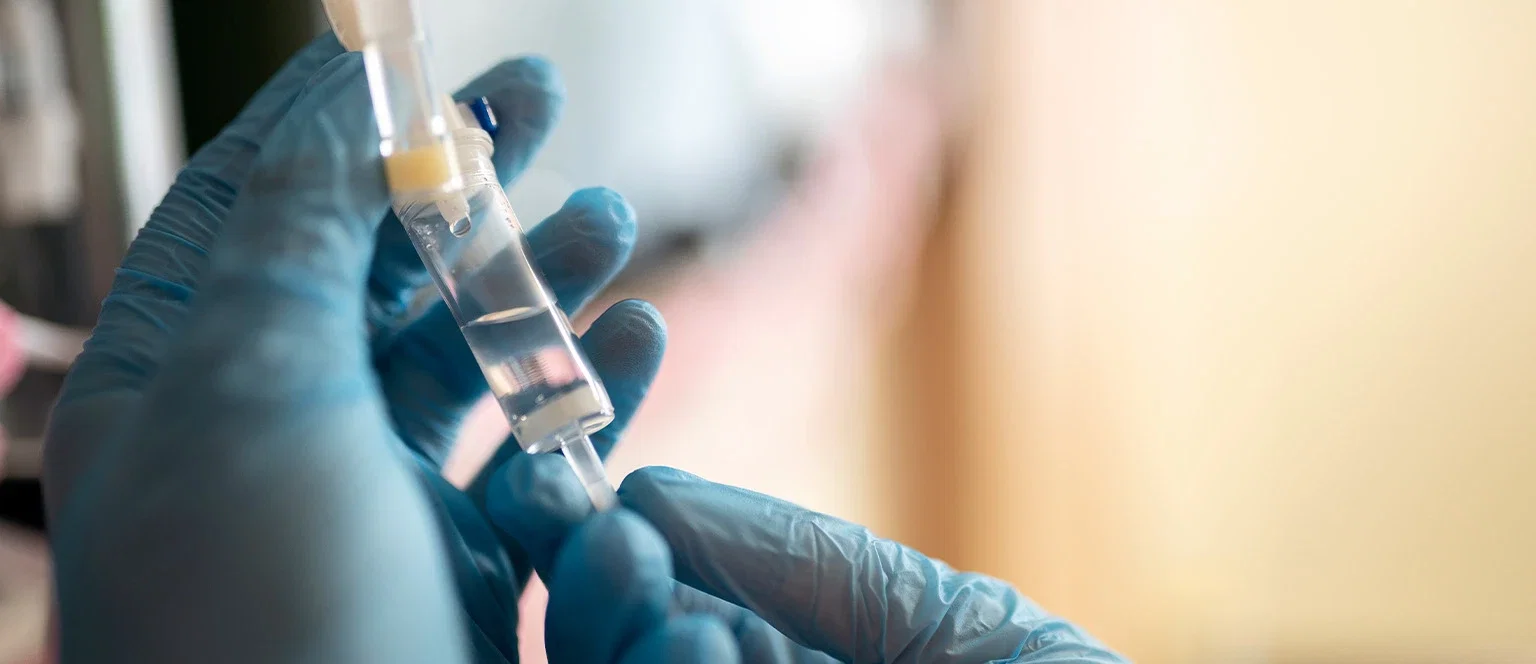 Close-up of gloved hands in blue nitrile gloves handling a small medical vial connected to an IV line