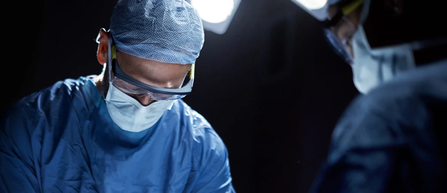 Surgeon in scrubs mask and safety glasses focused under bright operating room lights during a procedure
