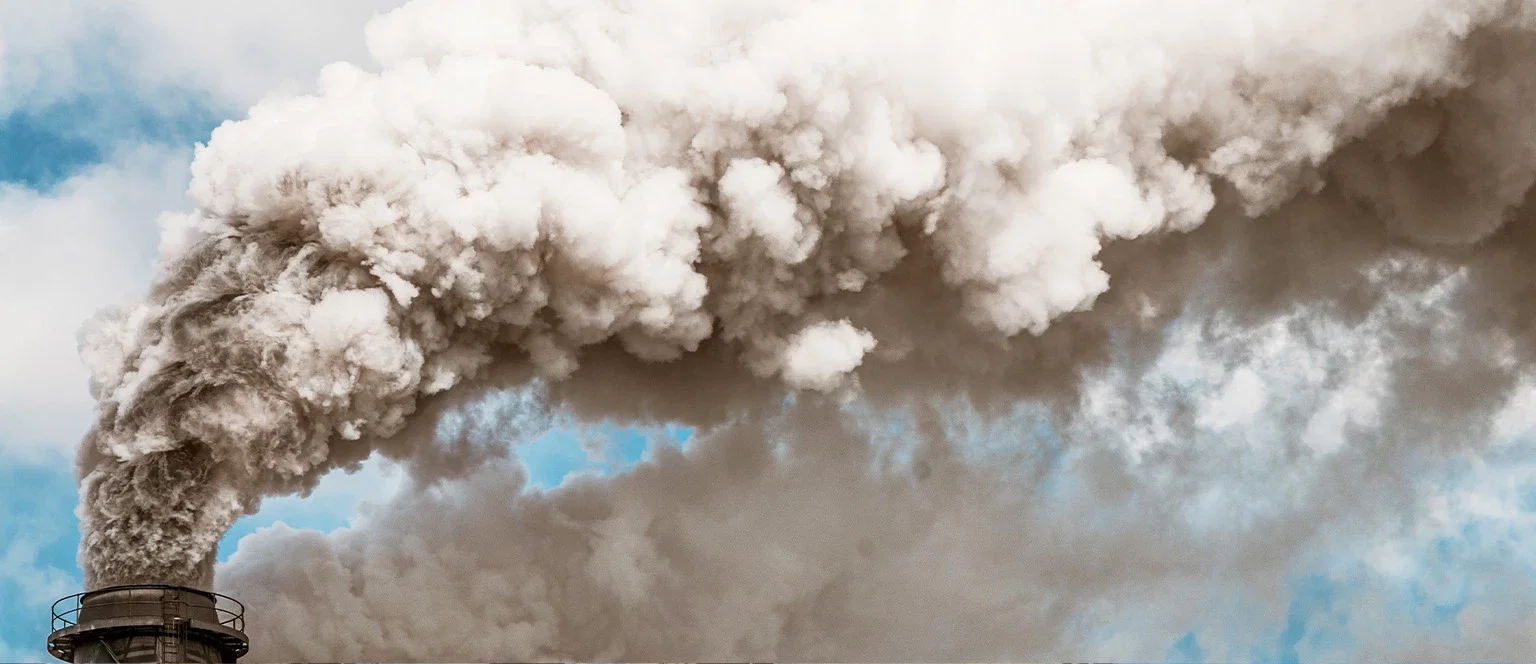 Dense gray smoke billowing from an industrial smokestack against a partly cloudy blue sky