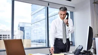Joel Feller in a dress shirt and tie standing at his desk, reviewing documents while talking on a phone, with a city view through large windows behind him