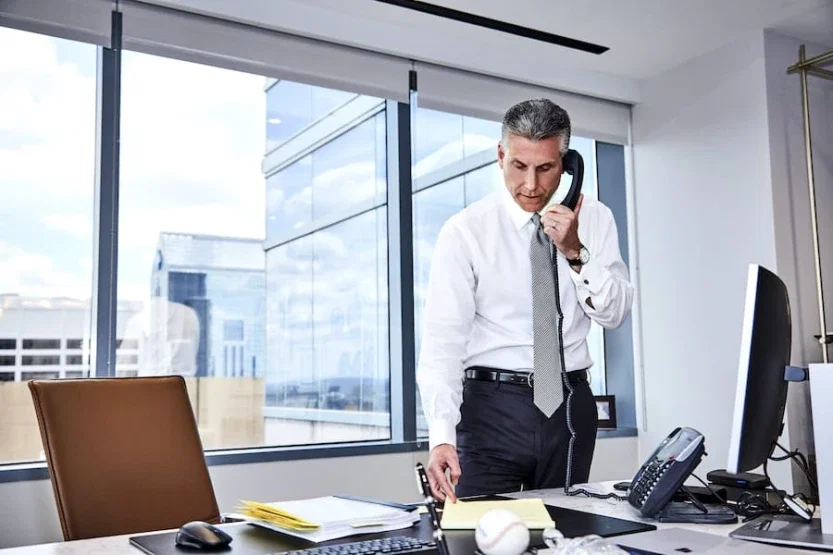 Joel Feller in a dress shirt and tie standing at his desk, reviewing documents while talking on a phone, with a city view through large windows behind him