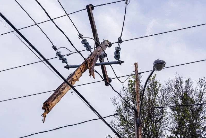A damaged wooden utility pole crossarm, split and leaning at an angle, with power lines and insulators tangled against an overcast sky