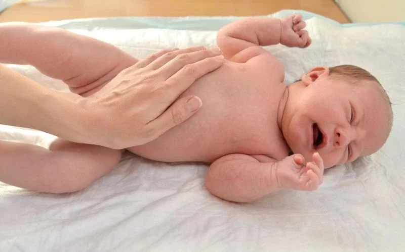 A crying newborn lying on a white surface while an adult hand gently supports the baby
