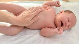 A crying newborn lying on a white surface while an adult hand gently supports the baby