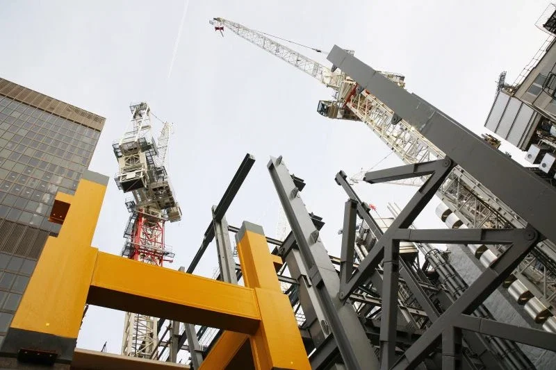 Low upward-angle view of a large active construction site, showing yellow and gray structural steel beams and two tall tower cranes against an overcast sky