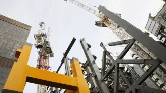 Low upward-angle view of a large active construction site, showing yellow and gray structural steel beams and two tall tower cranes against an overcast sky