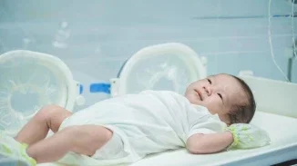 A newborn baby in a white onesie lying awake in a hospital incubator, looking upward