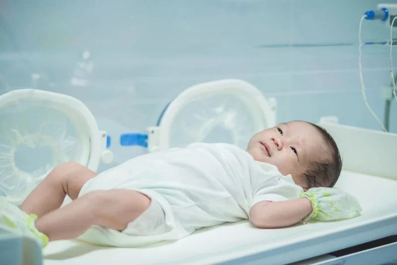 A newborn baby in a white onesie lying awake in a hospital incubator, looking upward