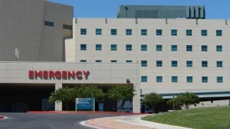 Exterior of a hospital emergency department entrance with a large red "Emergency" sign above the covered ambulance bay, under a clear blue sky