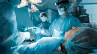 A patient lying on an operating table wearing a surgical cap, with a surgical team in blue scrubs and masks working around her under bright operating lights