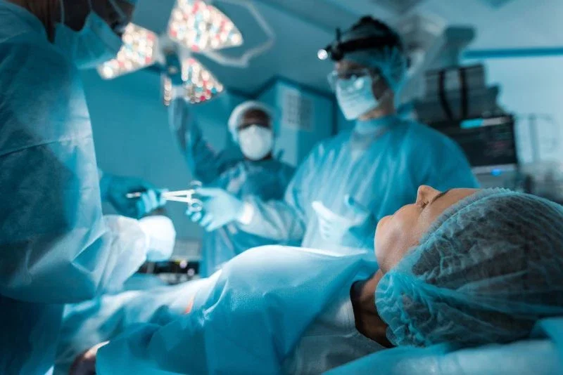 A patient lying on an operating table wearing a surgical cap, with a surgical team in blue scrubs and masks working around her under bright operating lights