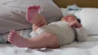 A newborn baby lying on its back on a white surface with legs raised, wearing a light gray onesie