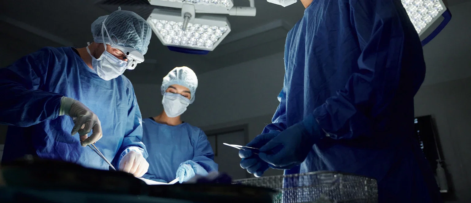 A surgical team in blue scrubs, caps, masks, and gloves performing an operation under bright overhead lights in an operating room